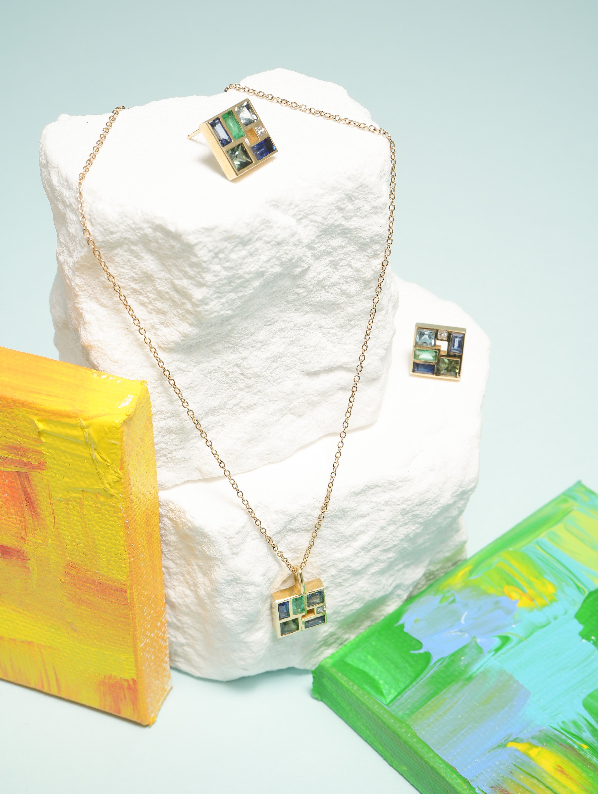 Gold necklace with pendant on a textured white block, with abstract art pieces in the background.